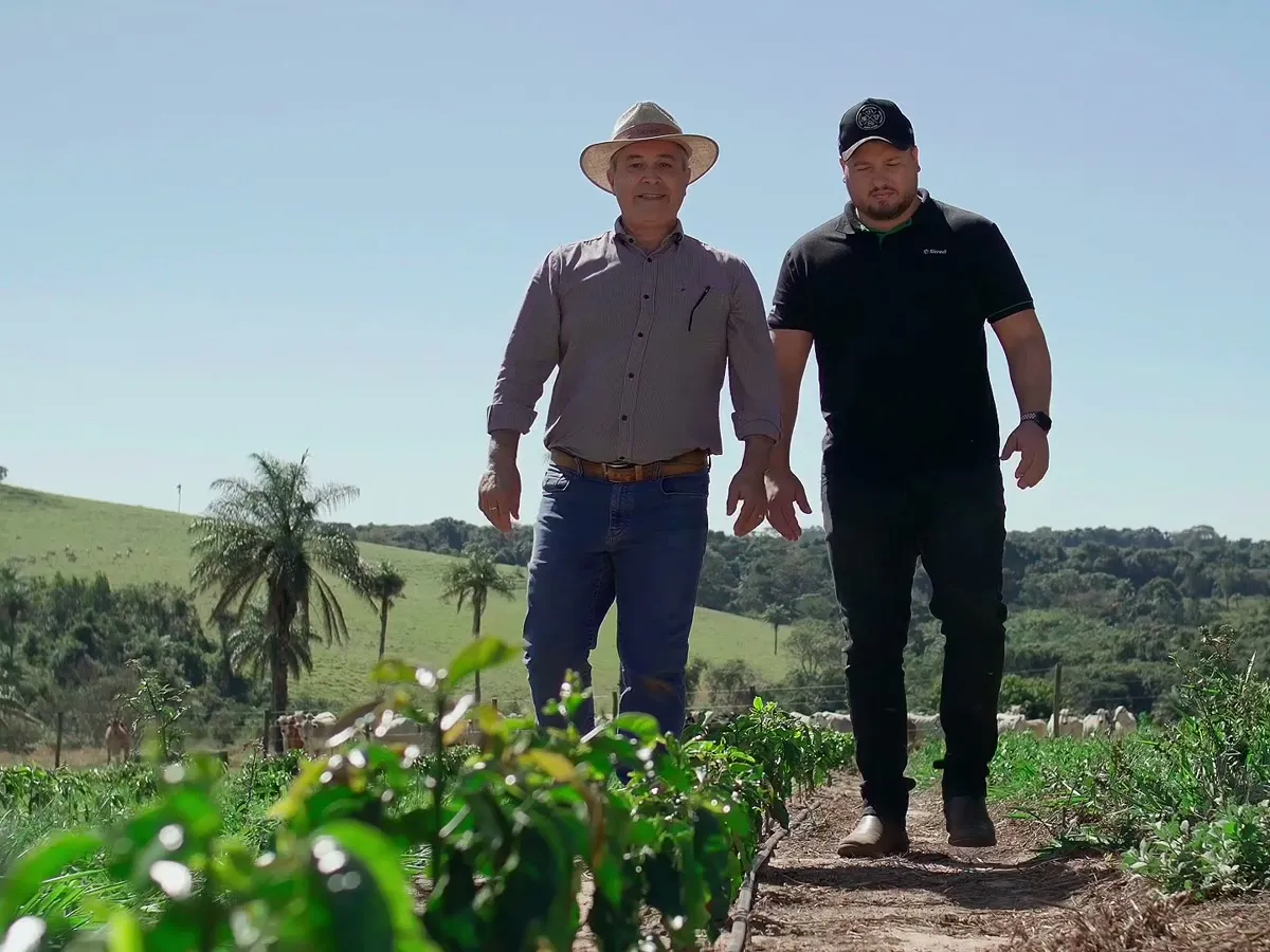 Dois homens caminham lado a lado por uma lavoura, com morros ao fundo